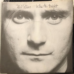 Peut inclure: Pochette d'album en noir et blanc avec un gros plan sur le visage d'un homme. Le texte manuscrit en haut indique "Phil Collins" et "In the Air tonight". L'image est un portrait de l'artiste.