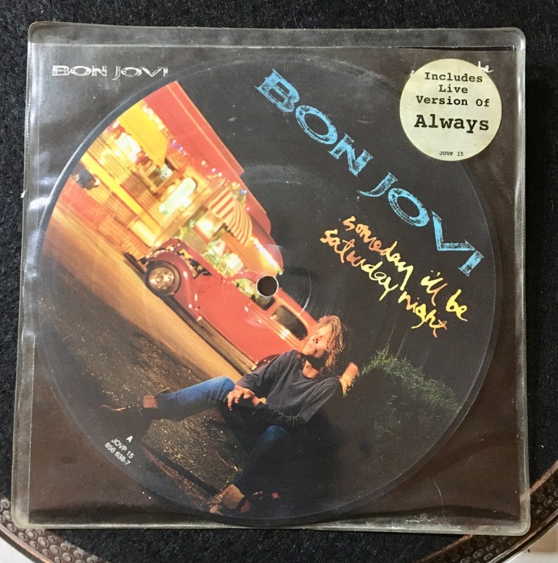 May include: A black and white vinyl record with a picture of a man sitting on a curb in front of a building. The record is titled "Someday I'll Be Saturday Night" by Bon Jovi. The record includes a live version of the song "Always".