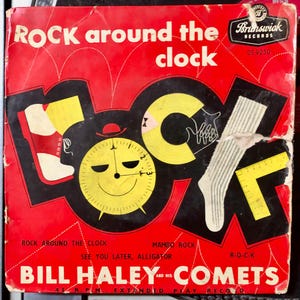 May include: A red vinyl record cover with a black and yellow illustration of a clock, a shoe, and a sock. The text on the cover reads "Rock around the clock" and "Bill Haley and His Comets".