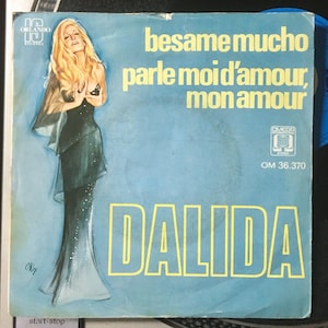 May include: A vintage vinyl record cover featuring a woman in a black dress with a blue background. The title of the record is "besame mucho parle moi d'amour, mon amour" and the artist is "DALIDA". The record label is "OMEGA INTERNATIONAL" and the catalog number is "OM 36.370".