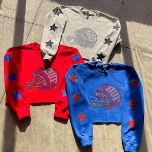 May include: Three cropped sweatshirts with long sleeves and star designs on the sleeves. The sweatshirts are in red, blue, and beige. Each sweatshirt has a rhinestone football helmet with the word "BUF" on it.