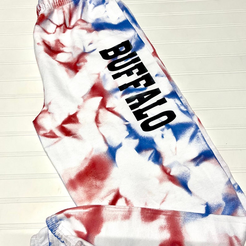 Może przedstawiać: Białe spodnie dresowe z wzorem tie-dye w kolorze czerwonym i niebieskim. Słowo "BUFFALO" jest nadrukowane na nogawce czarnym kolorem.