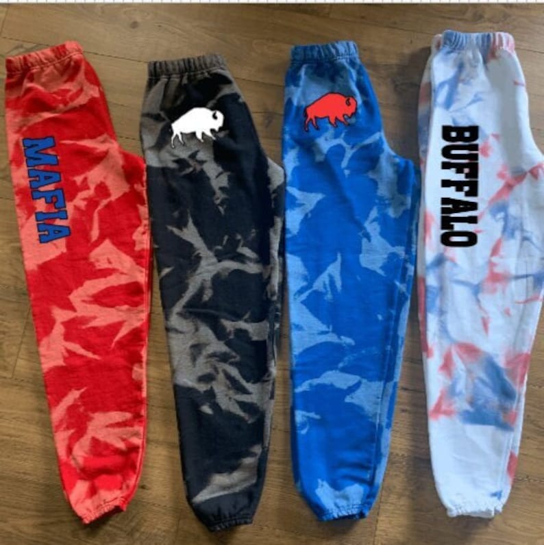 Może przedstawiać: Cztery pary dres&oacute;w tie-dye z r&oacute;żnymi wzorami i tekstem. Dresy są czerwone, niebieskie, czarne i białe z wzorami tie-dye. Tekst na dresach brzmi "MAFIA", "BUFFALO", sylwetka bawoła i sylwetka bizona.