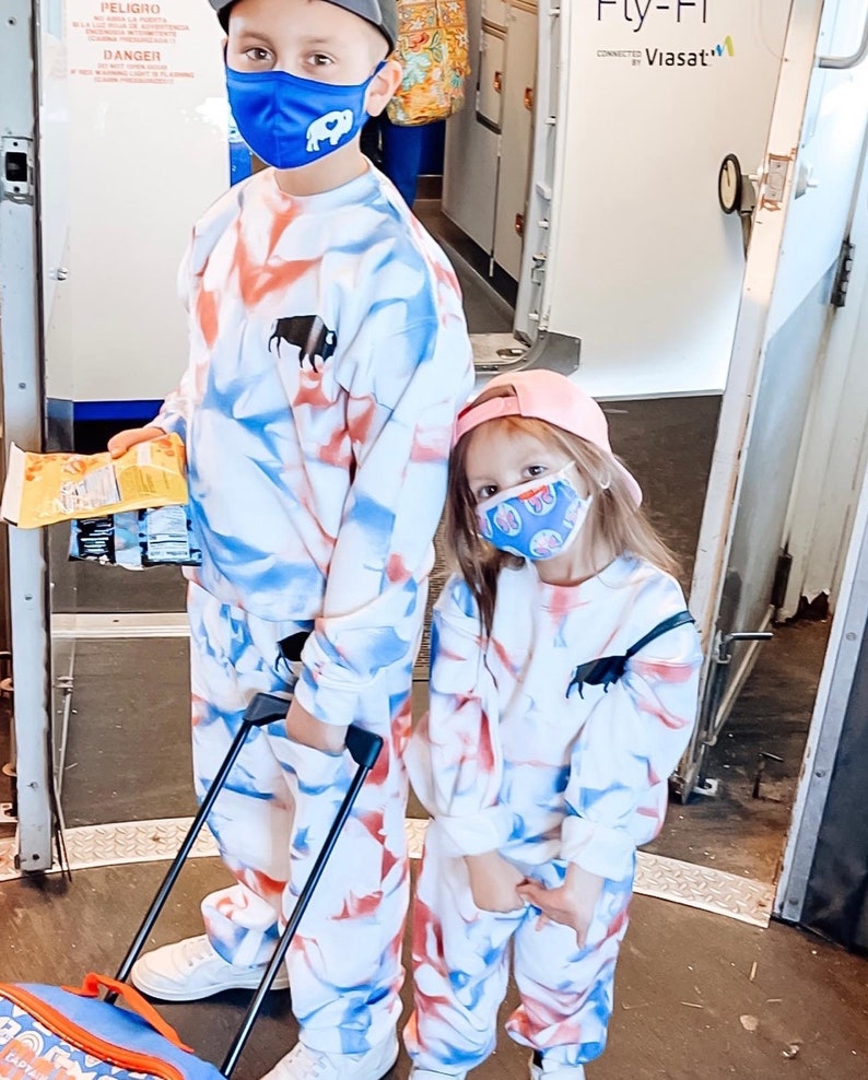 Może przedstawiać: Dwoje dzieci w dopasowanych dresach w kolorze białym, niebieskim i czerwonym z nadrukiem tie-dye i czarnym logo bawoła. Dzieci mają na sobie niebieskie i wzorzyste maseczki. Chłopiec trzyma worek chips&oacute;w i walizkę na k&oacute;łkach. Dziewczynka ma na sobie r&oacute;żowy kapelusz. W tle widoczny jest napis "Fly-Fi Connected by Viasat".