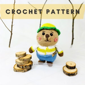 Puede incluir: Una muñeca de oso de ganchillo con un sombrero amarillo y verde, una camisa blanca con rayas amarillas y pantalones azules. La muñeca sostiene un pequeño palo de madera. El texto "CROCHET PATTERN" está en la parte superior de la imagen.