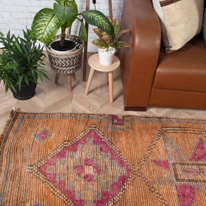 May include: An orange and pink patterned rug with a geometric design. The rug has a distressed look with visible wear and tear. The rug is placed on a light-colored wooden floor. Plants and a leather sofa are in the background.