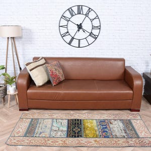 May include: A brown leather sofa with two decorative pillows sits against a white brick wall. A large black clock hangs above the sofa. A colorful rug is on the floor in front of the sofa.