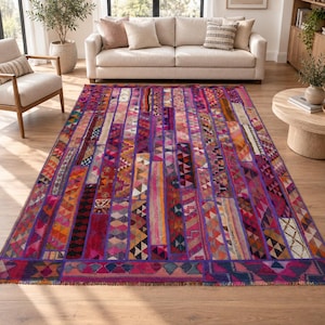 Puede incluir: Una alfombra rectangular con un diseño de patchwork vibrante compuesto por tiras verticales en tonos magenta, morado, naranja y rosa. La alfombra muestra varios patrones geométricos, incluyendo rombos, triángulos y zigzags, con un borde de flecos.