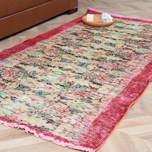 Peut inclure: Un tapis rectangulaire au fond beige orné d'un motif répétitif de fleurs noires, vertes et roses. Le tapis est encadré par une large bordure rouge unie et présente des franges courtes de couleur rose clair sur les extrémités.