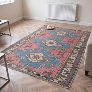 May include: A rectangular area rug featuring a vibrant pink and blue color palette with geometric patterns, including central diamond motifs and a decorative border with fringe edges, laid out on a light-colored wooden floor in a room.