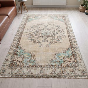 May include: A rectangular area rug featuring a beige field with a central medallion and ornate floral motifs in shades of light blue, brown, and cream. The border displays a repeating pattern of intricate designs in matching earth tones.