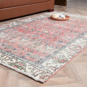 May include: A rectangular, distressed-look rug with a faded floral pattern in shades of red, blue, and cream. The rug has a cream-colored border with floral and leaf motifs. The rug is placed on a light-colored wooden floor.