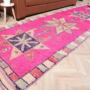 May include: A bright pink runner rug featuring geometric star designs in beige and purple. The rug has a patterned border in shades of pink, beige, and green. This decorative rug is suitable for hallways and entryways.