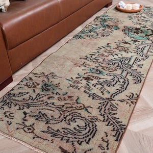 May include: A long, rectangular runner rug featuring a faded beige base with intricate, symmetrical floral and geometric patterns in shades of dark brown, muted green, and soft teal. The rug has a low-pile texture and short fringe at the ends.