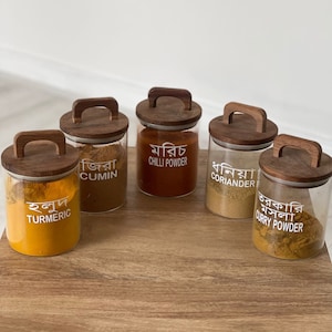 May include: Five glass spice jars with wooden lids and handles. The jars are filled with different spices and labelled in Bengali: turmeric, cumin, chilli powder, coriander, and curry powder.