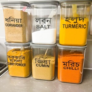 May include: Six clear plastic containers with grey lids, each filled with a different spice. The containers are labelled with the English name of the spice and the Bengali name. The spices are coriander, salt, turmeric, curry powder, cumin, and chilli.