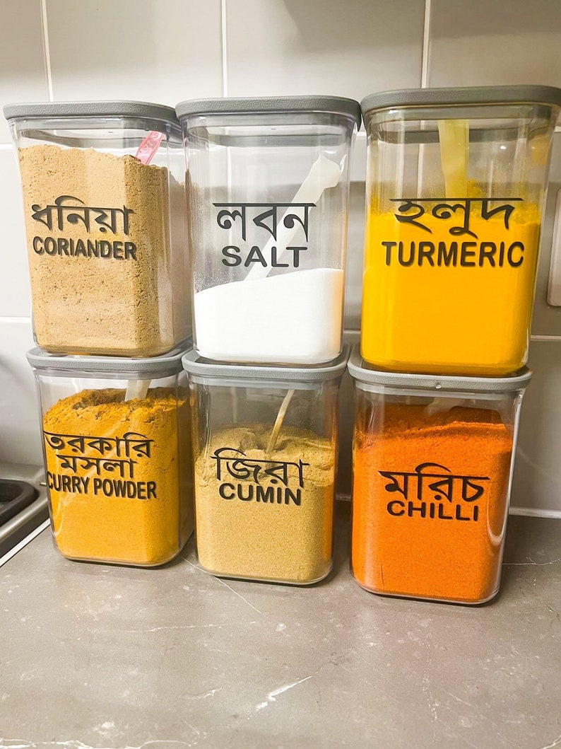 May include: Six clear plastic containers with grey lids, each filled with a different spice. The containers are labelled with the English name of the spice and the Bengali name. The spices are coriander, salt, turmeric, curry powder, cumin, and chilli.