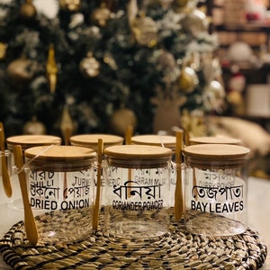 May include: Six clear glass jars with wooden lids and spoons. Each jar is labelled with a different spice in Bengali and English. The spices are: Chili, Dried Onion, Turmeric, Garam Masala, Coriander Powder, and Bay Leaves.