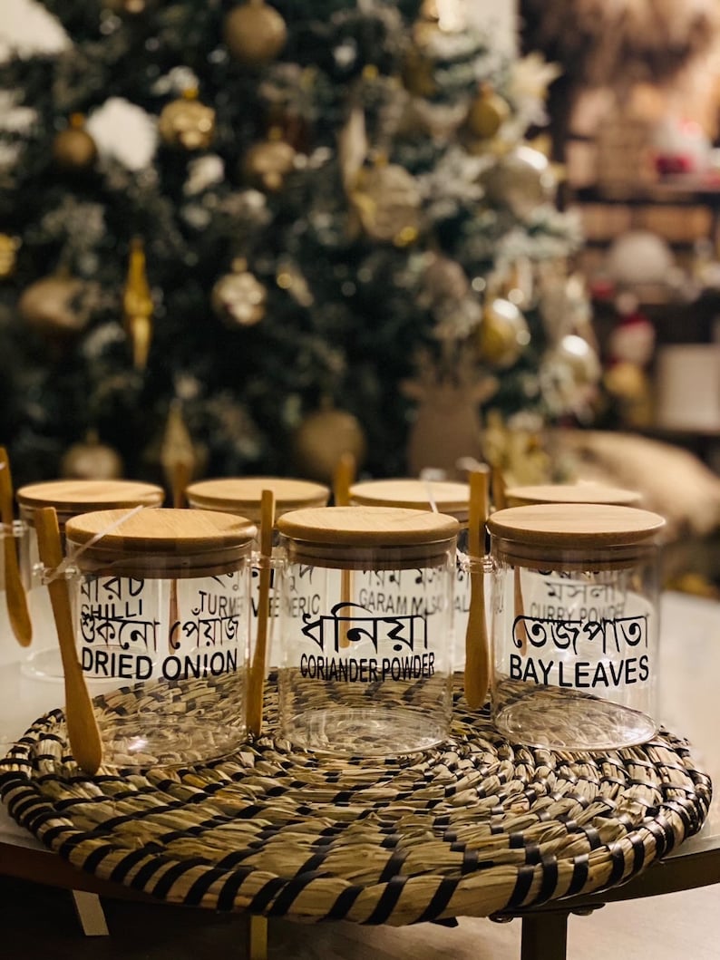 May include: Six clear glass jars with wooden lids and spoons. Each jar is labelled with a different spice in Bengali and English. The spices are: Chili, Dried Onion, Turmeric, Garam Masala, Coriander Powder, and Bay Leaves.