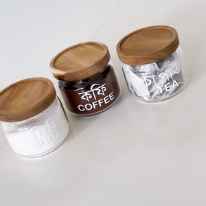 May include: Three glass jars with wooden lids, labelled "Sugar", "Coffee" and "Tea". The jars are filled with white sugar, brown coffee grounds, and tea bags.