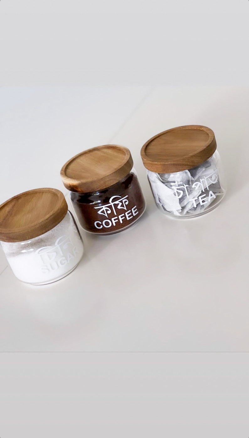 May include: Three glass jars with wooden lids, labelled "Sugar", "Coffee" and "Tea". The jars are filled with white sugar, brown coffee grounds, and tea bags.