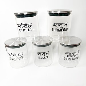 May include: Five glass spice jars with metal lids. The jars are labelled with the names of spices in both English and Bengali. The spices are: Chilli, Turmeric, Coriander Powder, Salt, and Curry Powder.
