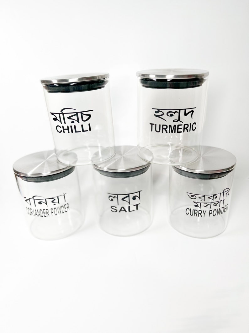 May include: Five glass spice jars with metal lids. The jars are labelled with the names of spices in both English and Bengali. The spices are: Chilli, Turmeric, Coriander Powder, Salt, and Curry Powder.