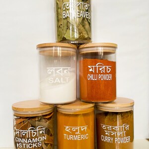 May include: Six glass jars with wooden lids, each containing different spices. The jars are stacked in a pyramid shape. The labels on the jars are in Bengali and English, and they read: Bay Leaves, Salt, Chilli Powder, Cinnamon Sticks, Turmeric, and Curry Powder.