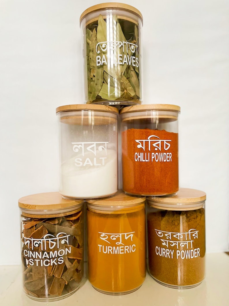 May include: Six glass jars with wooden lids, each containing different spices. The jars are stacked in a pyramid shape. The labels on the jars are in Bengali and English, and they read: Bay Leaves, Salt, Chilli Powder, Cinnamon Sticks, Turmeric, and Curry Powder.