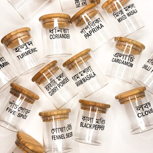 May include: A set of 14 glass spice jars with bamboo lids. Each jar has a black label with the name of the spice written in Bengali. The spices include turmeric, coriander, paprika, mixed masala, curry powder, garam masala, cardamom, chilli, five spice, fennel seed, black pepper, and clove.