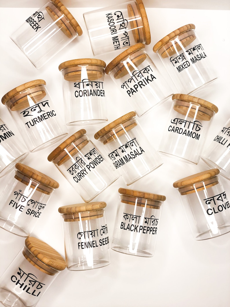May include: A set of 14 glass spice jars with bamboo lids. Each jar has a black label with the name of the spice written in Bengali. The spices include turmeric, coriander, paprika, mixed masala, curry powder, garam masala, cardamom, chilli, five spice, fennel seed, black pepper, and clove.