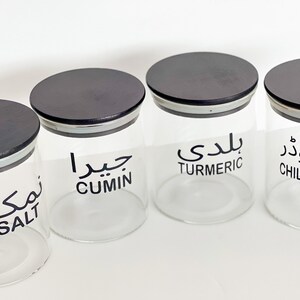 May include: Four glass spice jars with black lids and labels. The labels are in Arabic and English. The spices are salt, cumin, turmeric, and chilli powder.
