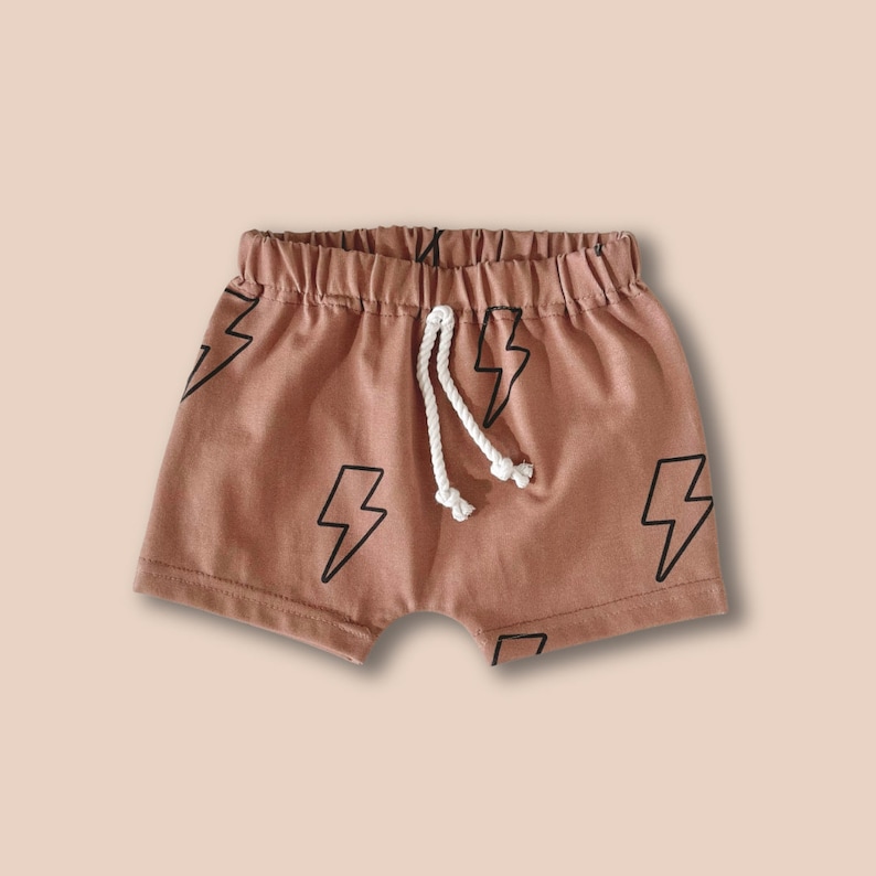 May include: Dusty rose shorts featuring a black lightning bolt pattern printed across the fabric. The shorts have an elasticized waistband with a white braided drawstring cord tied in the center, providing a secure fit for the garment.