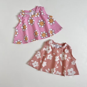 May include: Two sleeveless tops. One is light pink with a repeating pattern of smiling daisy faces. The other is a muted rose color with a white floral pattern. Both tops have a flared, A-line shape.