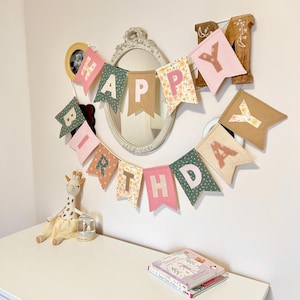 May include: A fabric birthday banner featuring individual pennants in shades of pink, green, and floral patterns, spelling out the words HAPPY BIRTHDAY in block letters. The banner is hung in a curved arrangement against a plain wall.