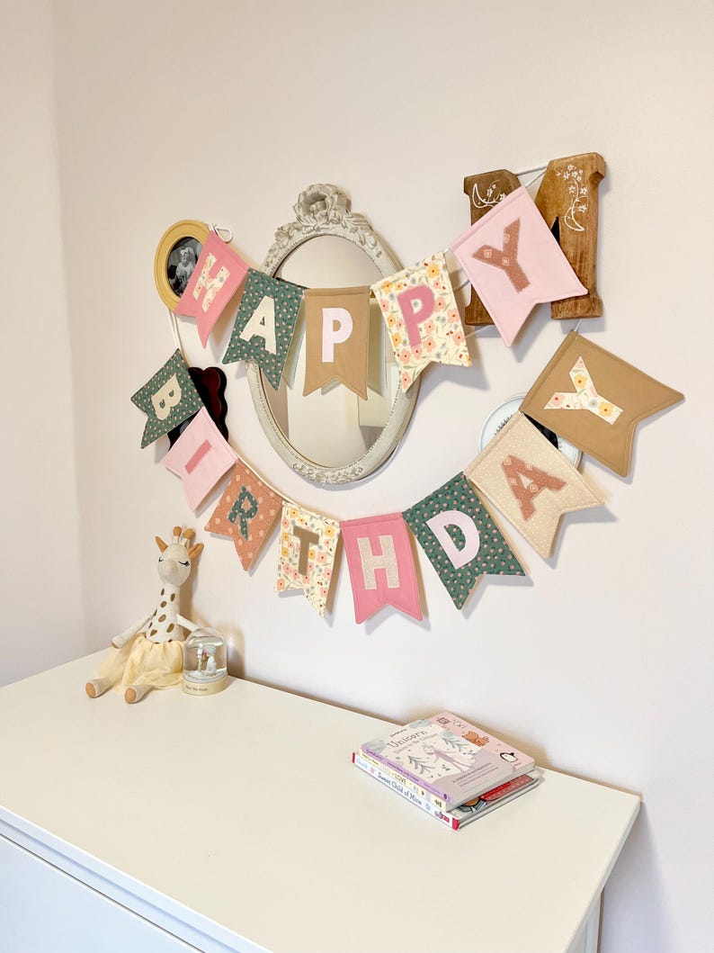 May include: A fabric birthday banner featuring individual pennants in shades of pink, green, and floral patterns, spelling out the words HAPPY BIRTHDAY in block letters. The banner is hung in a curved arrangement against a plain wall.