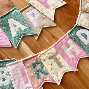 May include: A fabric banner featuring individual pennants with the letters spelling HAPPY BIRTHDAY. Each pennant is crafted from various cotton fabrics in shades of pink, green, beige, and floral patterns, with contrasting fabric letters appliqued on.