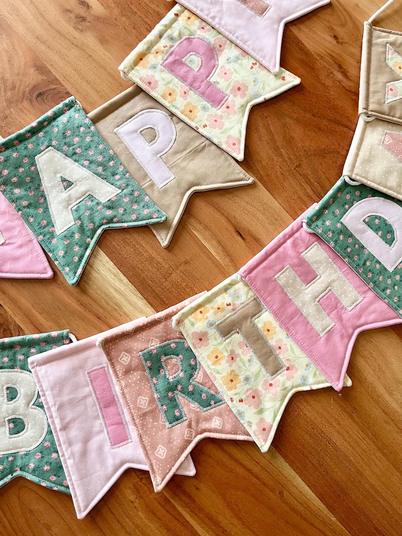 May include: A fabric banner featuring individual pennants with the letters spelling HAPPY BIRTHDAY. Each pennant is crafted from various cotton fabrics in shades of pink, green, beige, and floral patterns, with contrasting fabric letters appliqued on.