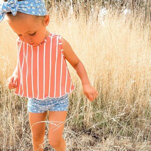 May include: A young child wearing a peach-colored, vertically striped top and blue floral shorts. A matching blue floral headband with a bow is in the child's hair. The child is wearing light green sandals and standing in tall, dry grass.