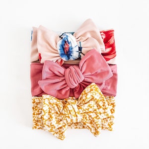 May include: Three fabric headbands with knotted bows. The top headband is pink with a floral print, the middle headband is a solid dusty rose color, and the bottom headband is yellow with a white floral print.