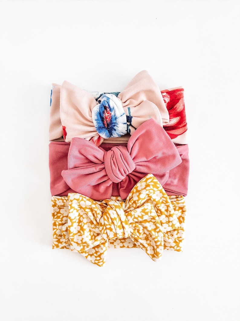 May include: Three fabric headbands with knotted bows. The top headband is pink with a floral print, the middle headband is a solid dusty rose color, and the bottom headband is yellow with a white floral print.