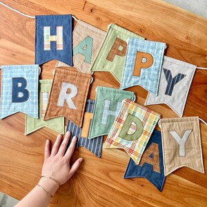 May include: A fabric banner spelling out HAPPY BIRTHDAY with individual swallowtail pennants. Each pennant features a unique combination of solid, plaid, and striped cotton fabrics in shades of blue, green, brown, and tan with appliqued letters.