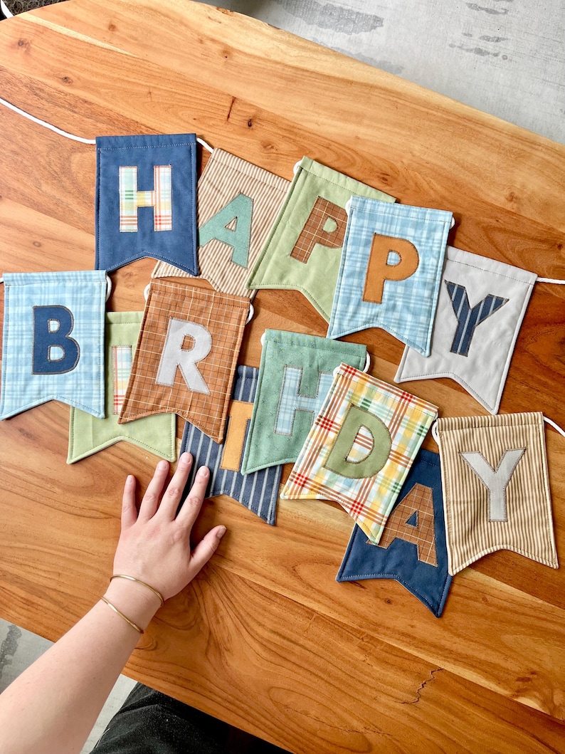 May include: A fabric banner spelling out HAPPY BIRTHDAY with individual swallowtail pennants. Each pennant features a unique combination of solid, plaid, and striped cotton fabrics in shades of blue, green, brown, and tan with appliqued letters.