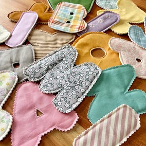May include: Assortment of colorful fabric letters in various patterns and colors, including pink, green, yellow, and floral prints. The letters are cut with a scalloped edge and appear to be handmade, possibly for crafting or educational purposes.