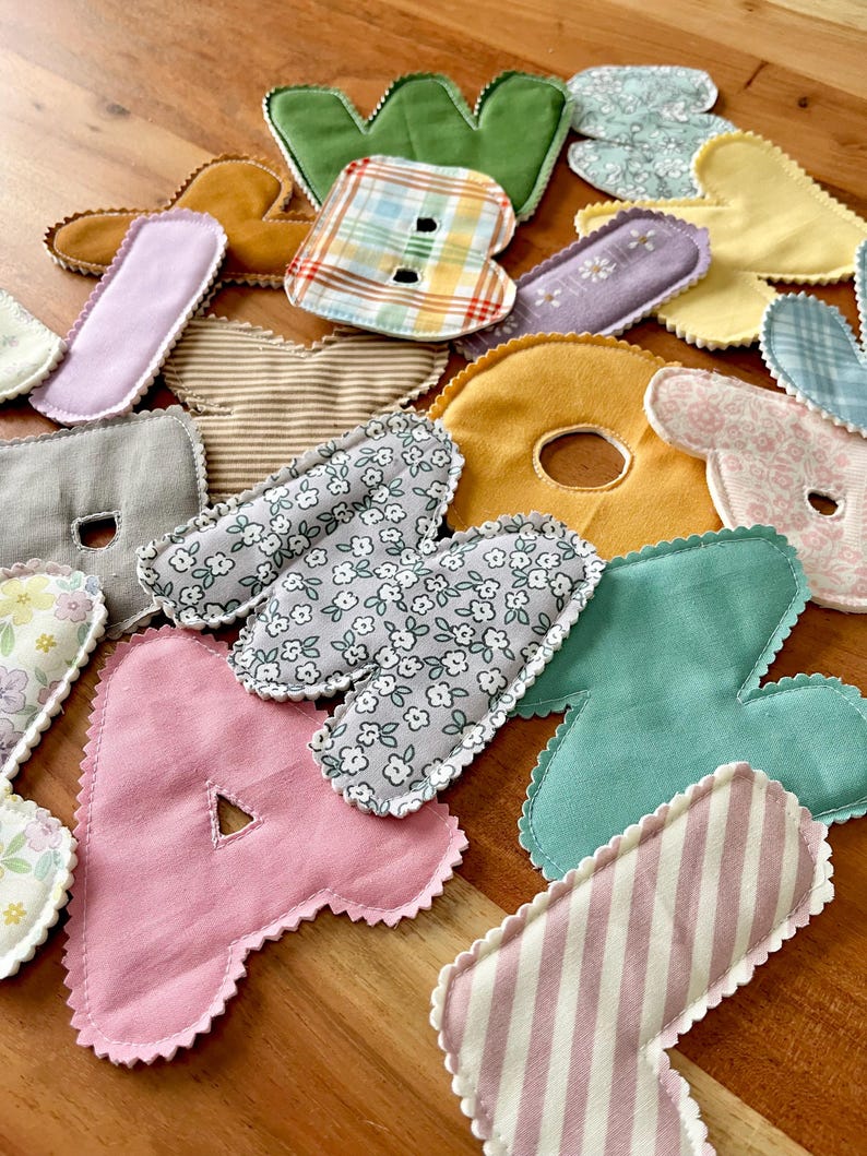May include: Assortment of colorful fabric letters in various patterns and colors, including pink, green, yellow, and floral prints. The letters are cut with a scalloped edge and appear to be handmade, possibly for crafting or educational purposes.