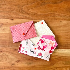 Op de afbeelding: Een verzameling handgemaakte stoffen envelopportemonnees in verschillende patronen. E&eacute;n is roze met witte stippen, een andere is cr&egrave;me met een bloemenpatroon en de derde heeft harten, rozen en het woord "lovely". Elke portemonnee heeft een drukknoopsluiting.