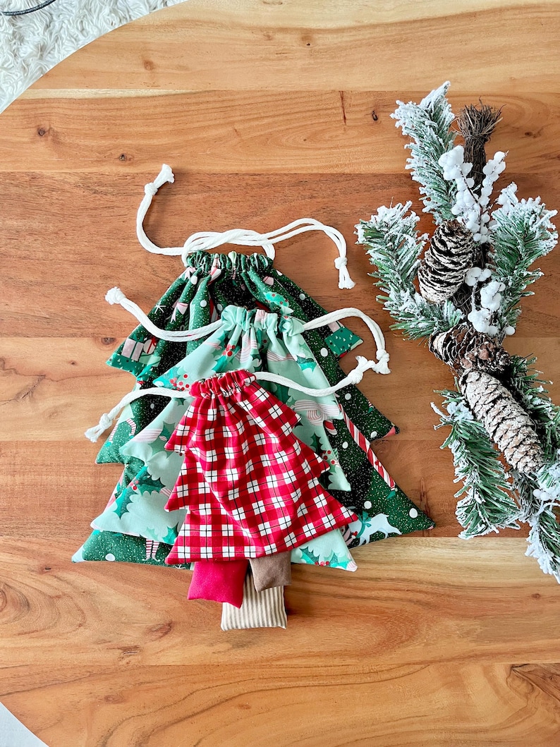 May include: A Christmas tree-shaped collection of fabric gift bags. The bags are green, red, and white, with various holiday patterns. Each bag has a drawstring closure. A festive pine branch with snow and pinecones is in the background.