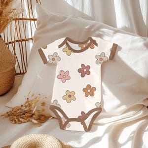 K&ouml;nnte beinhalten: Ein wei&szlig;er Baby-Strampler mit braunem Besatz und einem Muster aus bunten Blumen. Der Strampler hat kurze &Auml;rmel und einen Druckknopfverschluss am unteren Ende.