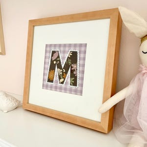 May include: A square wooden frame containing a white mat and a central fabric letter M. The letter features a dark brown background with a floral pattern in shades of orange, cream, and green, finished with a white scalloped stitched border.