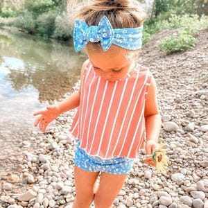 May include: A young child wearing a blue floral headband with a matching bow, a coral and white striped top, and blue floral shorts. The child is holding a yellow flower and standing on a rocky shore.